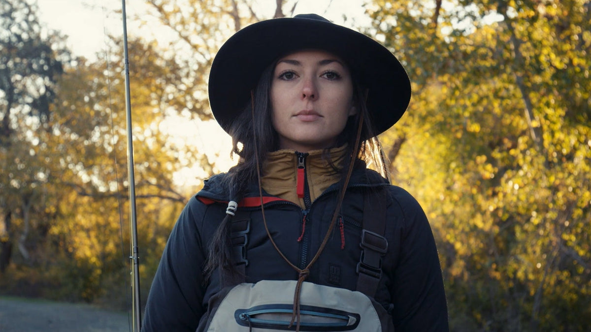 黄葉した木々を背に、つば広の帽子をかぶりフライロッドを持って立つKayla Lockhart